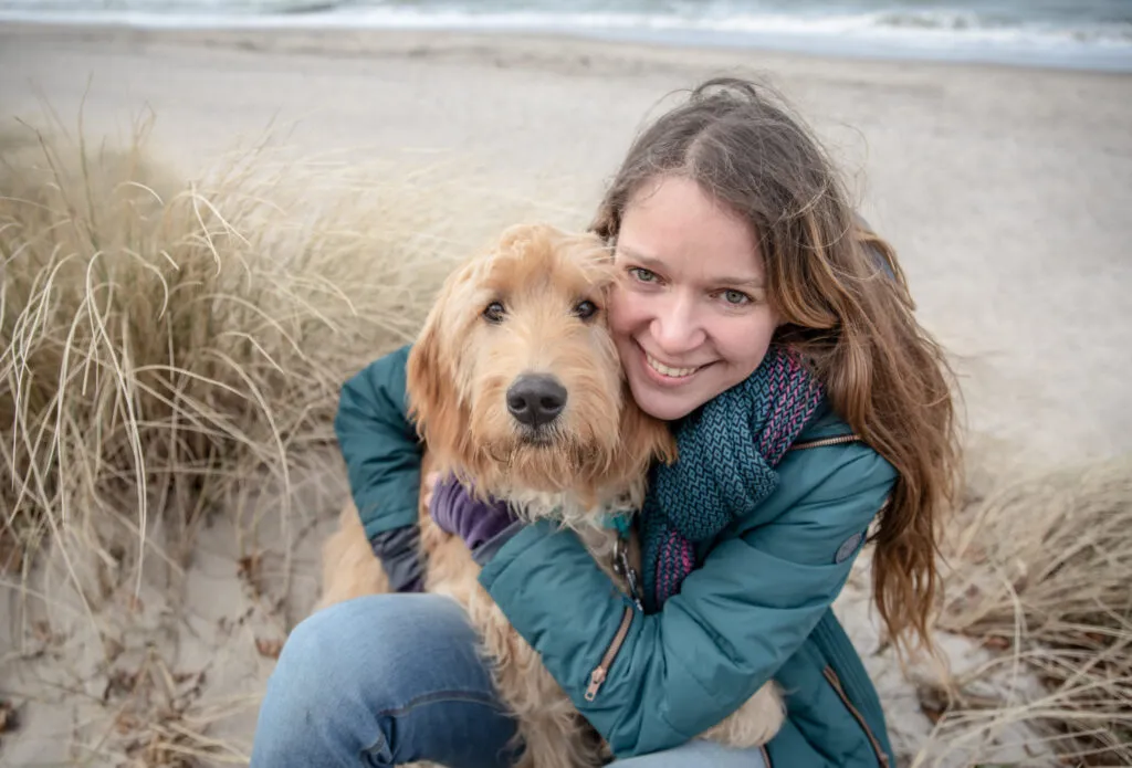 Eine Frau mit langen braunen Haaren lächelt, während sie einen großen, flauschigen Hund umarmt. Sie sind draußen an einem Sandstrand mit hohem Gras. Im Hintergrund ist das Meer unter einem bewölkten Himmel zu sehen.